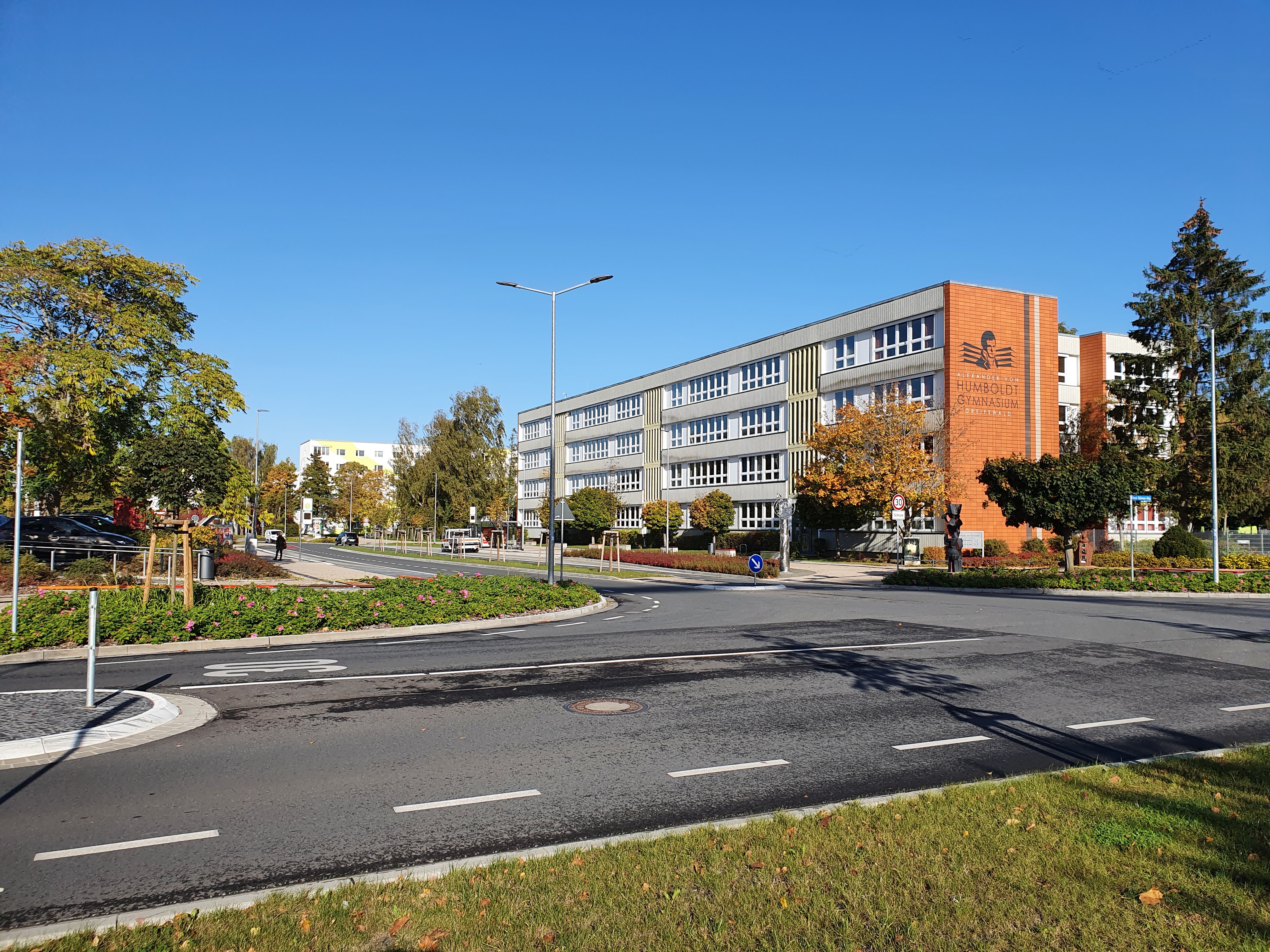 Ein langes Schulgebäude mit einigen Bäumen davor und im Vordergrund eine Kreuzung