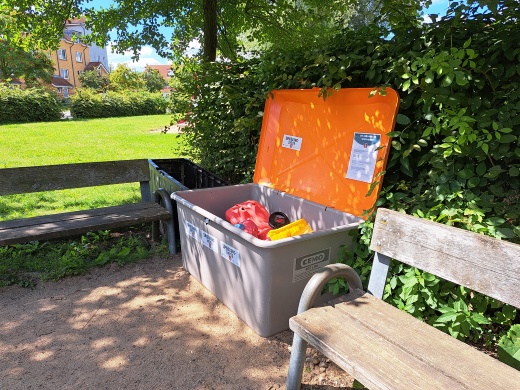 Spielzeugkiste im Poeler Weg © Quartierskoordination der Universitäts- und Hansestadt Greifswald Eine Kiste mit einem orangefarbenen Deckel gefüllt mit Spielzeug zwischen zwei Bänken auf einem Spielplatz