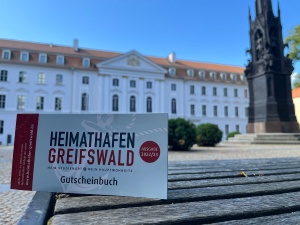 Foto des Gutscheinbuches Heimathafen Greifswald 2022 vor dem Universitätshauptgebäude - © Theres Behnke Foto des Gutscheinbuches Heimathafen Greifswald 2022 vor dem Universitätshauptgebäude