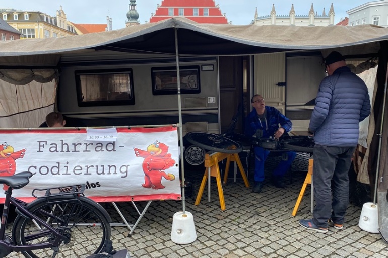 Ein Mitarbeiter der ABS unterhält sich mit einem Bürger in seinem Zelt, vor sich das Fahrrad eingespannt, um es zu codieren