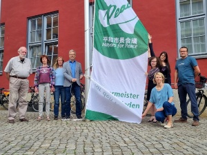 Mitglieder der Bürgerschaft, der Oberbürgermeister und Mitarbeitende des Beauftragtenbüros hissen der Mayors for Peace Flagge vor dem Rathaus