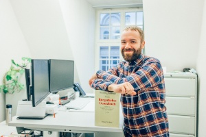 Ein Auszubildender steht vor seinem Büroschreibtisch und lehnt sich mit den Armen auf ein Gesetzbuch - © Philipp Marthaler Ein Auszubildender steht vor seinem Büroschreibtisch und lehnt sich mit den Armen auf ein Gesetzbuch