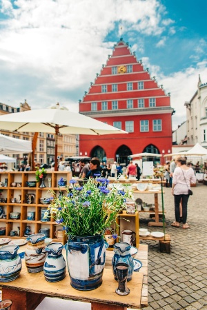 Auf dem Historischen Marktplatz findet ein Sondermarkt statt. Im Vordergrund ist ein Frühlingsstrauß Blumen und ein Töpferstand zu sehen, im Hintergrund schlendern Gäste über den Markt.