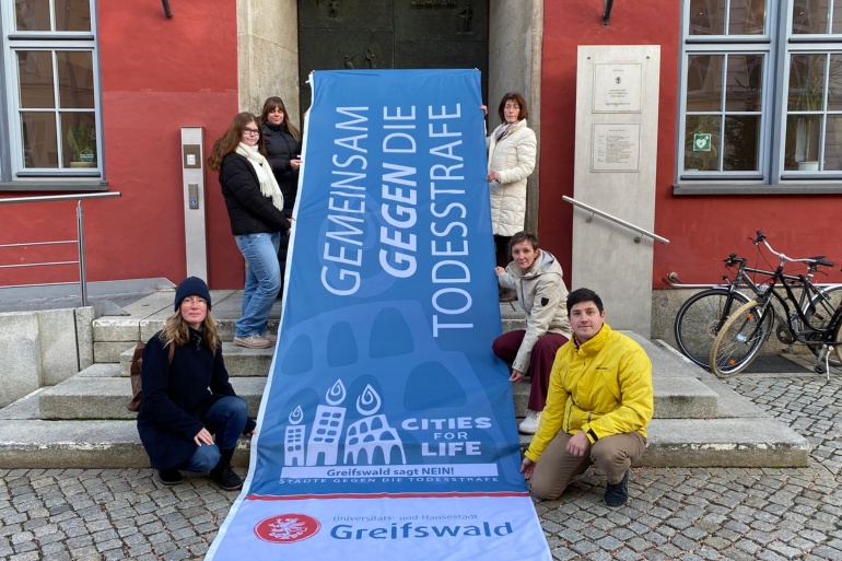 Mitarbeitende mit der Flagge des Bündnisses Städte gegen die Todesstrafe vor dem Rathaus