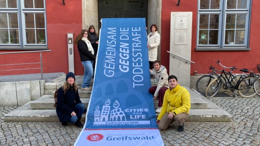 Mitarbeitende mit der Flagge des Bündnisses Städte gegen die Todesstrafe vor dem Rathaus