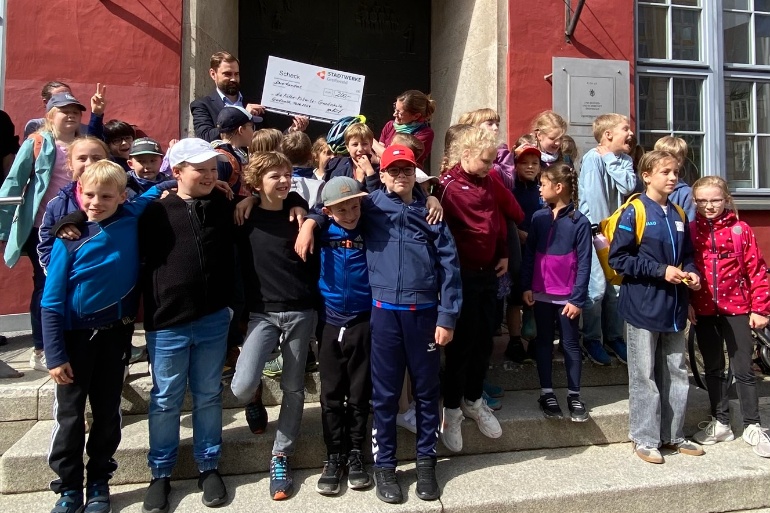 Kinder der Käthe-Kollwitz-Grundschule stehen auf der Treppe vor dem Rathaus und halten einen 200-Euro-Gutschein der Stadtwerke in die Luft